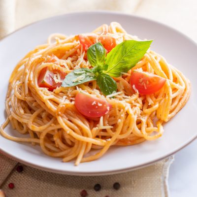 Tasty appetizing classic italian spaghetti pasta with tomato sauce, cheese parmesan and basil on plate and ingredients for cooking pasta on white marble table.